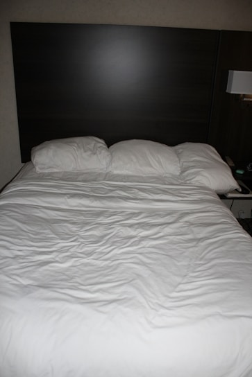 A neatly made bed with three pillows resting against a dark wooden headboard. The bedspread is white and appears slightly wrinkled. Beside the bed, there is a wall-mounted lamp and a portion of a bedside table visible.