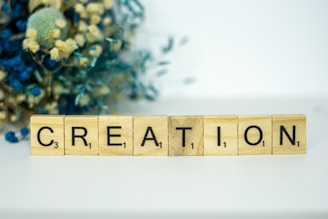 Wooden Scrabble tiles arranged to spell the word 'CREATION' are placed on a white surface. In the background, there is a blurred arrangement of dried flowers, including blue and beige hues, adding a touch of elegance.