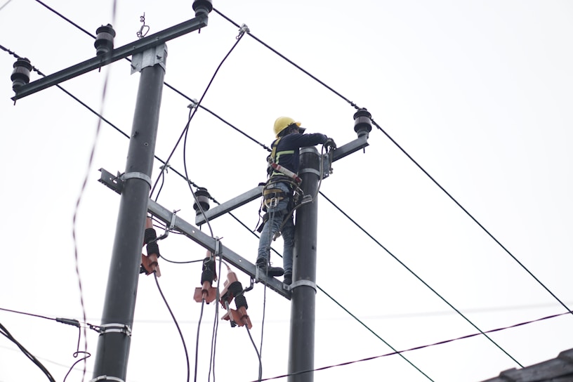 A person wearing a safety harness and helmet is working high on an electrical pole surrounded by wires.