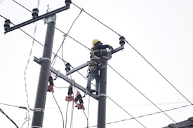 A person wearing a safety harness and helmet is working high on an electrical pole surrounded by wires.