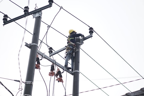 A person wearing a safety harness and helmet is working high on an electrical pole surrounded by wires.