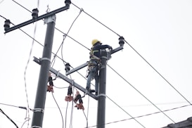 A person wearing a safety harness and helmet is working high on an electrical pole surrounded by wires.