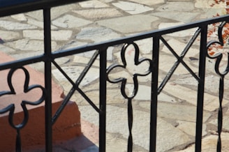 An ornate black metal railing with floral and geometric patterns in the foreground, set against a background of a light stone patio.