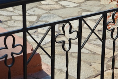 An ornate black metal railing with floral and geometric patterns in the foreground, set against a background of a light stone patio.