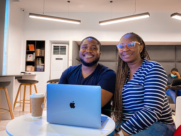 Two people are seated side by side at a round table with a laptop and a coffee cup in a modern, well-lit room. The room has a minimalist design with bookshelves, bar stools, and an air conditioning unit in the background. Both individuals are smiling, conveying a sense of friendliness and comfort.