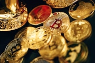 A collection of shiny, metallic coins featuring various cryptocurrency symbols such as Bitcoin, Ethereum, and others, scattered on a surface. The coins are primarily golden in color with intricate designs. Some coins have embossed logos and text, reflecting the lighting to create a polished and luxurious appearance.