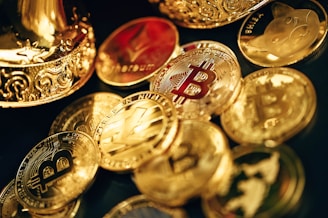 A collection of shiny, metallic coins featuring various cryptocurrency symbols such as Bitcoin, Ethereum, and others, scattered on a surface. The coins are primarily golden in color with intricate designs. Some coins have embossed logos and text, reflecting the lighting to create a polished and luxurious appearance.