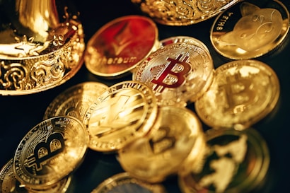A collection of shiny, metallic coins featuring various cryptocurrency symbols such as Bitcoin, Ethereum, and others, scattered on a surface. The coins are primarily golden in color with intricate designs. Some coins have embossed logos and text, reflecting the lighting to create a polished and luxurious appearance.