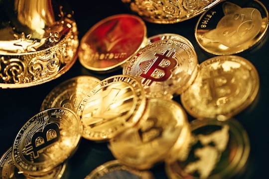 A collection of shiny, metallic coins featuring various cryptocurrency symbols such as Bitcoin, Ethereum, and others, scattered on a surface. The coins are primarily golden in color with intricate designs. Some coins have embossed logos and text, reflecting the lighting to create a polished and luxurious appearance.