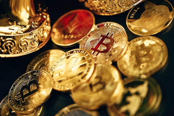 A collection of shiny, metallic coins featuring various cryptocurrency symbols such as Bitcoin, Ethereum, and others, scattered on a surface. The coins are primarily golden in color with intricate designs. Some coins have embossed logos and text, reflecting the lighting to create a polished and luxurious appearance.
