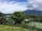 A lush landscape with a large mountain in the background, partially covered by clouds. The foreground features a variety of greenery, including trees and shrubs, and a tennis court is visible among the vegetation. The sky is partly cloudy with patches of blue.