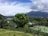 A lush landscape with a large mountain in the background, partially covered by clouds. The foreground features a variety of greenery, including trees and shrubs, and a tennis court is visible among the vegetation. The sky is partly cloudy with patches of blue.