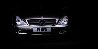 A sleek, silver luxury car is set against a dark background, with its headlights illuminating the front grille. The prominent emblem on the hood indicates a high-end brand, and the license plate is visible beneath the grille.