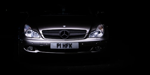 A sleek, silver luxury car is set against a dark background, with its headlights illuminating the front grille. The prominent emblem on the hood indicates a high-end brand, and the license plate is visible beneath the grille.