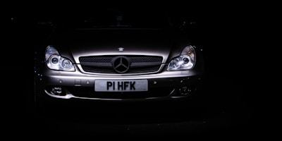 A sleek, silver luxury car is set against a dark background, with its headlights illuminating the front grille. The prominent emblem on the hood indicates a high-end brand, and the license plate is visible beneath the grille.