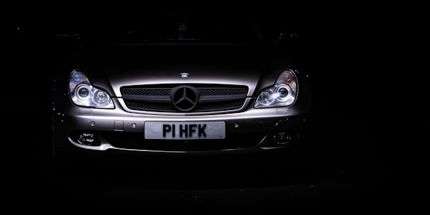 A sleek, silver luxury car is set against a dark background, with its headlights illuminating the front grille. The prominent emblem on the hood indicates a high-end brand, and the license plate is visible beneath the grille.
