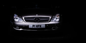 A sleek, silver luxury car is set against a dark background, with its headlights illuminating the front grille. The prominent emblem on the hood indicates a high-end brand, and the license plate is visible beneath the grille.