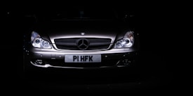 A sleek, silver luxury car is set against a dark background, with its headlights illuminating the front grille. The prominent emblem on the hood indicates a high-end brand, and the license plate is visible beneath the grille.