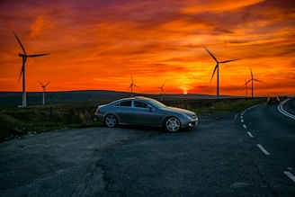 A silver car is parked on the side of a road at sunset with several large wind turbines in the background. The sky is filled with dramatic orange and yellow hues, creating a striking contrast with the landscape. The road curves away into the distance, where another vehicle can be seen driving away.