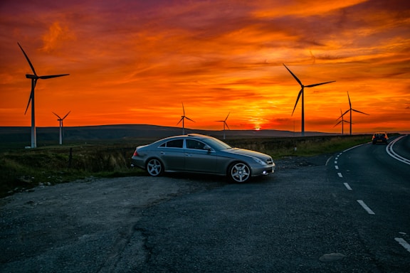 A silver car is parked on the side of a road at sunset with several large wind turbines in the background. The sky is filled with dramatic orange and yellow hues, creating a striking contrast with the landscape. The road curves away into the distance, where another vehicle can be seen driving away.