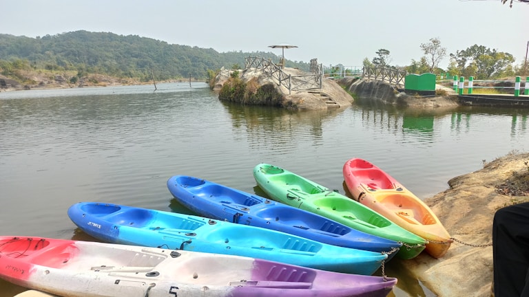 Colorful kayaks are lined up on the edge of a tranquil lake, surrounded by lush greenery and a rocky embankment. A small bridge-like structure is visible in the background, leading to a slightly elevated area.