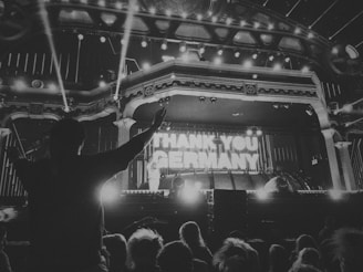 A black and white image of a crowded concert or event with prominent stage lighting and the phrase 'THANK YOU GERMANY' displayed at the center of the stage. A silhouetted figure with raised hands appears in the foreground, surrounded by an audience.