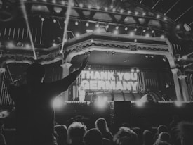 A black and white image of a crowded concert or event with prominent stage lighting and the phrase 'THANK YOU GERMANY' displayed at the center of the stage. A silhouetted figure with raised hands appears in the foreground, surrounded by an audience.