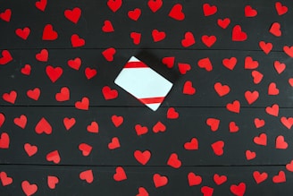 A black wooden surface is scattered with numerous red heart-shaped cutouts. In the center sits a small white gift box adorned with diagonal red stripes.