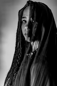 A person with braided hair partially covered by a sheer cloth. The lighting creates dramatic shadows across the face and fabric, adding depth and mystery.