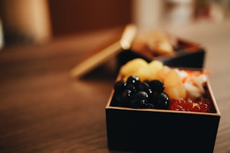 A close-up of a bento box containing a variety of foods including dark-colored beans, yellow fruit pieces, possibly pineapple, and small orange fish eggs. The focus is on the assortment within the box, which sits on a wooden surface with a soft, blurred background.