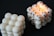 Two cube-shaped candles made from multiple wax spheres are set against a dark background. One candle is plain white, while the other features black spots on the spheres and is lit at the top.