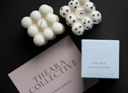 On a dark background, there are two uniquely shaped candles. One candle resembles a cluster of small spheres with a single wick at the center, while the other has a similar form but includes black dot patterns on the surface. Next to the candles, there is a card and a box, both displaying the name 'Theara Collective'.