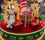 A festive Christmas display featuring two polar bear figures wearing red scarves, surrounded by large decorative gifts wrapped in shiny red and gold paper. Behind them are illuminated reindeer figures adorned with golden lights. The scene is set on a platform decorated with red and green fabric and bordered by glowing lights. A large candy cane and a Christmas tree are also visible in the background.
