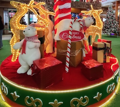 A festive Christmas display featuring two polar bear figures wearing red scarves, surrounded by large decorative gifts wrapped in shiny red and gold paper. Behind them are illuminated reindeer figures adorned with golden lights. The scene is set on a platform decorated with red and green fabric and bordered by glowing lights. A large candy cane and a Christmas tree are also visible in the background.