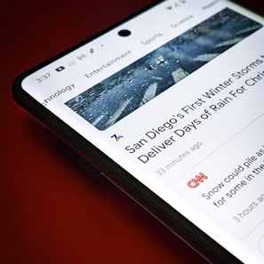 A smartphone screen displays a news article about San Diego's first winter storms, with a header image showing a wet road. The article is from a news app interface with sections like Technology, Entertainment, and Sports visible at the top. The background is a solid red color.