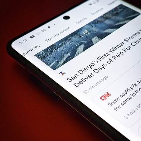 A smartphone screen displays a news article about San Diego's first winter storms, with a header image showing a wet road. The article is from a news app interface with sections like Technology, Entertainment, and Sports visible at the top. The background is a solid red color.