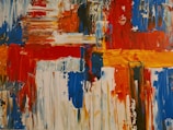 Bold, abstract art featuring vibrant blocks and strokes of color, including red, blue, orange, and white. The painting consists of thick, textured brushstrokes and dynamic patterns that evoke movement and energy.