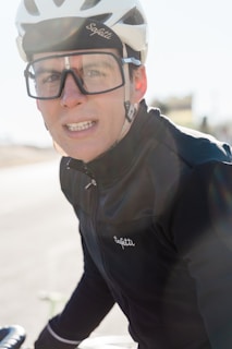 A person wearing a white cycling helmet and large glasses with a focused expression. They are dressed in a dark jacket branded 'Sopetti' and appear to be outdoors.