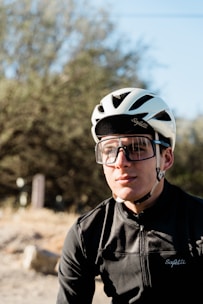 A young cyclist is wearing a white helmet and large protective sunglasses. He is dressed in a black cycling jersey with the brand 'Safetti' visible. The background features blurred greenery and a sunny sky.