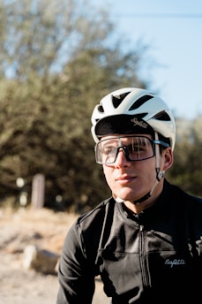 A young cyclist is wearing a white helmet and large protective sunglasses. He is dressed in a black cycling jersey with the brand 'Safetti' visible. The background features blurred greenery and a sunny sky.