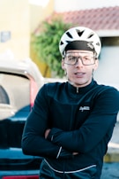 A person wearing a white cycling helmet and large protective glasses is standing with folded arms. They are wearing a black Safetti cycling jacket. In the background, a blurred view of a vehicle and a building with a sloped roof is visible.