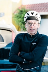 A person wearing a white cycling helmet and large protective glasses is standing with folded arms. They are wearing a black Safetti cycling jacket. In the background, a blurred view of a vehicle and a building with a sloped roof is visible.