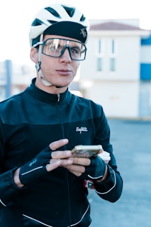 A cyclist is wearing a white helmet and large glasses, dressed in a black jacket with gloves. They are holding a smartphone and have a drink cup in hand. The background shows a blurry image of a building.