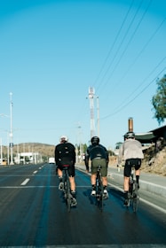 Three cyclists are riding on an open road, wearing helmets and athletic clothing. The sky is clear and blue with power lines visible running parallel to the road. There are hills and some buildings in the background, and one cyclist seems to be riding close to the roadside curb.