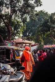 A person dressed in traditional orange attire is walking through a busy street. The person carries a bag and is near an auto-rickshaw. There are several people around, with some gathered near a vehicle. Tall trees and signs in an unknown language are visible in the background.