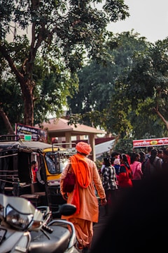 A person dressed in traditional orange attire is walking through a busy street. The person carries a bag and is near an auto-rickshaw. There are several people around, with some gathered near a vehicle. Tall trees and signs in an unknown language are visible in the background.