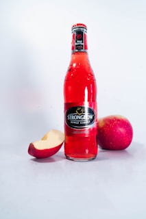A red bottle of Strongbow Apple Ciders with the label 'Red Berries' is placed upright on a light surface. Surrounding the bottle are two whole apples and a partially eaten apple slice.