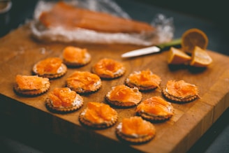 A wooden cutting board displays several crackers topped with smoked salmon and cream cheese. In the background, a knife rests near lemon slices, and a partially wrapped piece of smoked salmon is visible.