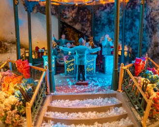 A miniature scene depicting an outdoor concert is set up in a gazebo. Small figures of musicians are positioned with instruments like a trumpet and trombone. The conductor stands with arms raised. The area is surrounded by colorful flowers and decorative elements, including red ribbons. Snow-like material covers the steps leading up to the stage.