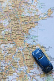 A detailed map of the Boston area with a small blue toy car placed on top of it. The map shows various cities and towns, highlighted roads and highways, and geographic features in the northeastern United States.